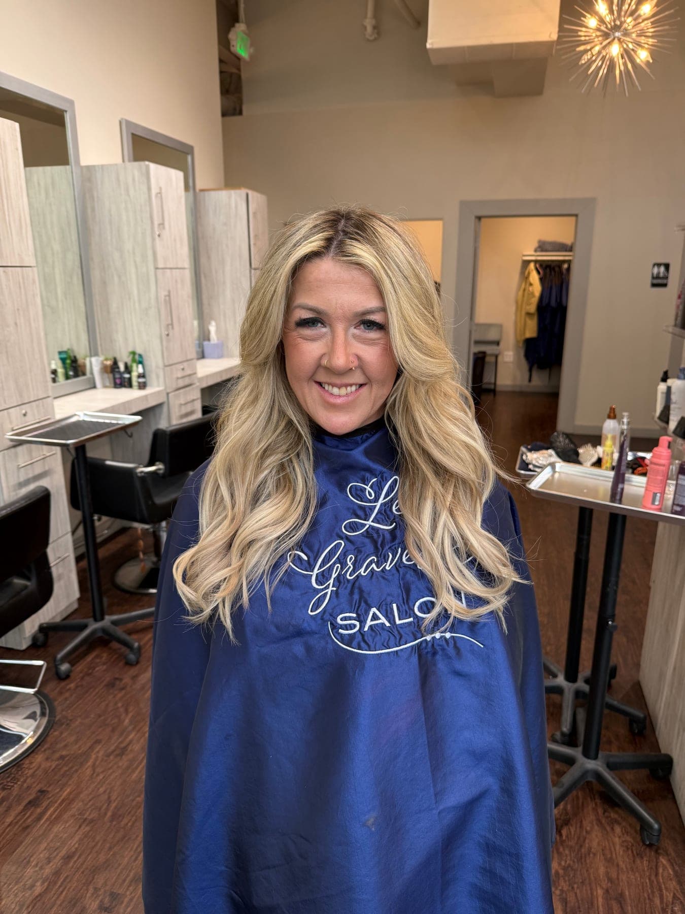 A client in a Lee Graves Salon cape on the salon floor in Plano, TX, with styling stations and chairs behind her